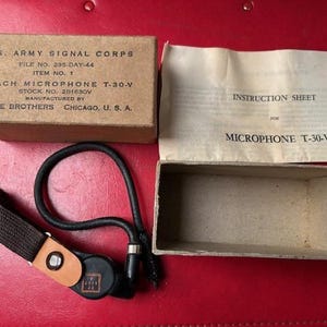Könnte beinhalten: Vintage U.S. Army Signal Corps Mikrofon T-30-V, mit Originalverpackung und Bedienungsanleitung. Die Box ist beige mit schwarzem Text, und das Mikrofon ist schwarz mit einem braunen Riemen. Das Anleitungsblatt ist beige.