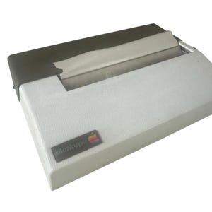 May include: A vintage Apple Silentype printer. The printer has a light gray body with a dark gray top. The Apple logo is visible on the front. The printer is designed to be compact and portable.