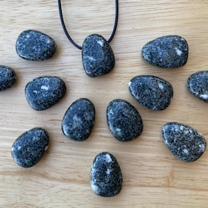 May include: A collection of dark gray, oval-shaped stones with white speckles, some arranged around a pendant on a black cord. The stones are displayed on a light brown wooden surface, creating a natural and earthy aesthetic.