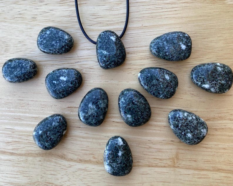May include: A collection of dark gray, oval-shaped stones with white speckles, some arranged around a pendant on a black cord. The stones are displayed on a light brown wooden surface, creating a natural and earthy aesthetic.