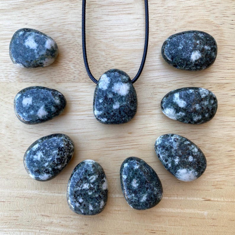 May include: A collection of dark green and white speckled stones, including a pendant on a black cord. The stones are oval and smooth, with a natural, organic appearance. The pendant is the focal point, surrounded by the other stones.
