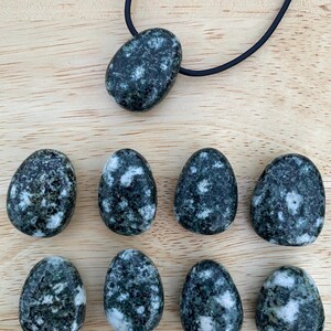 May include: A collection of dark green and white speckled stones, including a pendant on a black cord. The stones have an oval shape and are arranged on a light brown wooden surface. The pendant is a larger, similar stone.