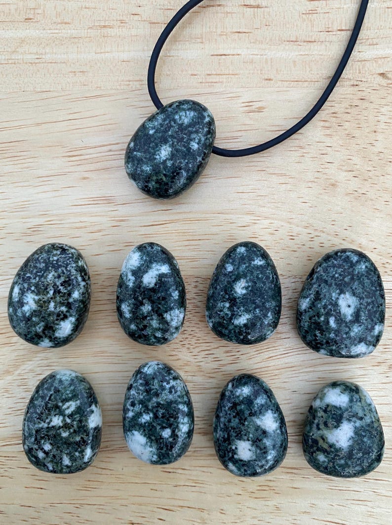May include: A collection of dark green and white speckled stones, including a pendant on a black cord. The stones have an oval shape and are arranged on a light brown wooden surface. The pendant is a larger, similar stone.