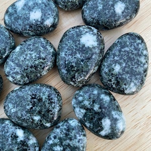 May include: A collection of tumbled stones with a speckled pattern of dark green and white. The stones are oval-shaped and vary slightly in size. They are arranged on a light-colored wooden surface.