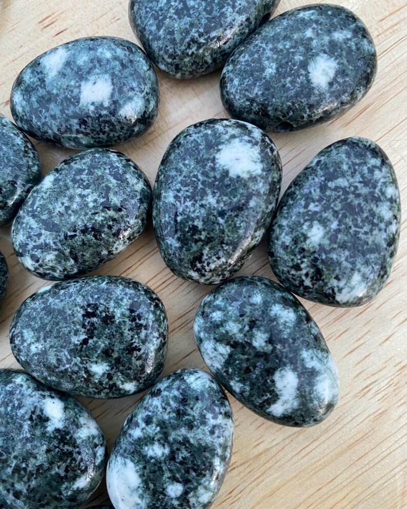 May include: A collection of tumbled stones with a speckled pattern of dark green and white. The stones are oval-shaped and vary slightly in size. They are arranged on a light-colored wooden surface.