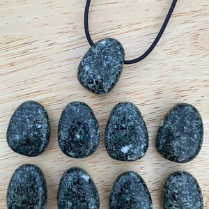 May include: A collection of dark green, speckled stones, some in an egg shape, and one pendant on a black cord. The stones have a mottled appearance with lighter green and white inclusions. The background is a light wood surface.