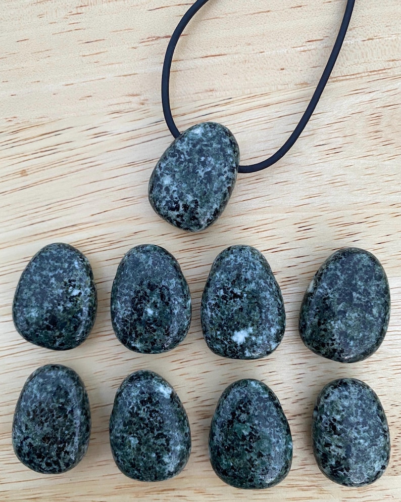 May include: A collection of dark green, speckled stones, some in an egg shape, and one pendant on a black cord. The stones have a mottled appearance with lighter green and white inclusions. The background is a light wood surface.