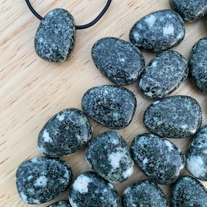 May include: A collection of polished, oval-shaped stones in shades of dark green and white, scattered on a wooden surface. One stone is suspended from a black cord, suggesting a pendant. The stones have a speckled appearance, with variations in color and texture.