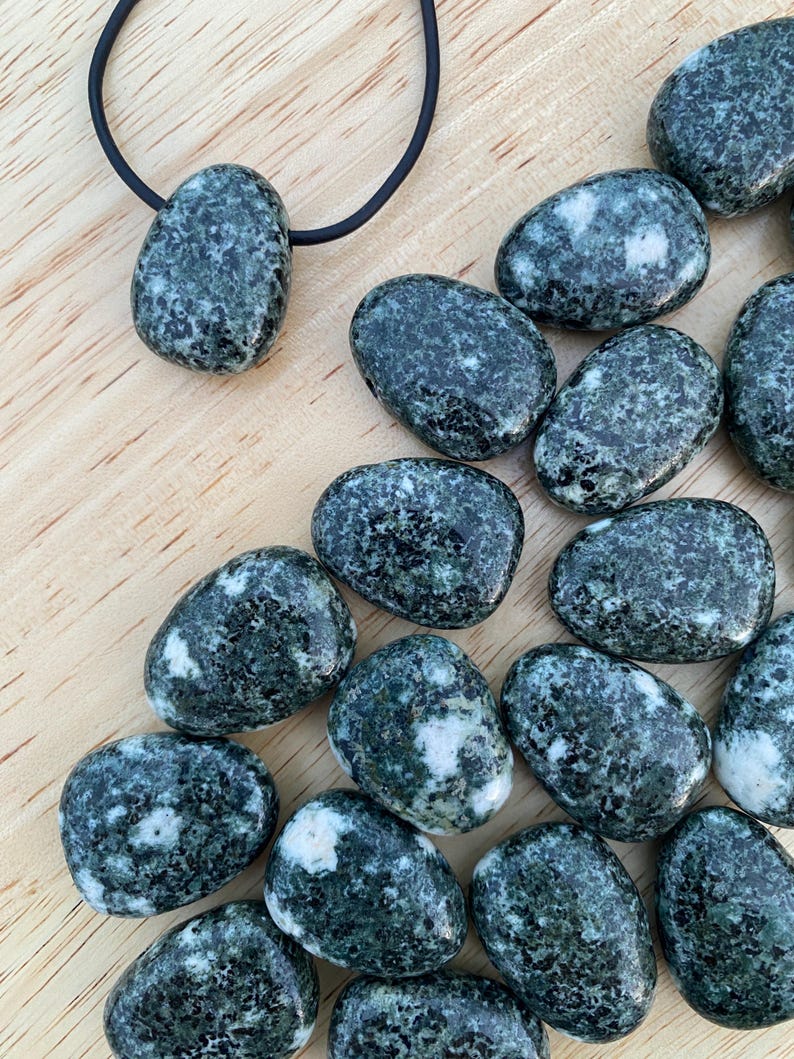 May include: A collection of polished, oval-shaped stones in shades of dark green and white, scattered on a wooden surface. One stone is suspended from a black cord, suggesting a pendant. The stones have a speckled appearance, with variations in color and texture.
