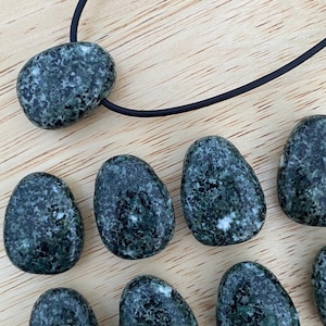 May include: A collection of dark green, oval-shaped stones with white speckles. One stone is strung on a black cord, forming a necklace. The stones are arranged on a light brown wooden surface.