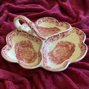 May include: A white, three-section serving dish with a handle. The dish has a scalloped edge and a red and white toile pattern featuring a building and floral border. The handle is white with a floral design.