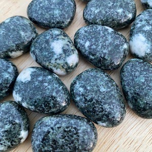 May include: A collection of smooth, oval-shaped stones. The stones are primarily dark green with white and light green marbling. The stones are arranged on a light brown wooden surface. The stones vary in size and shape.