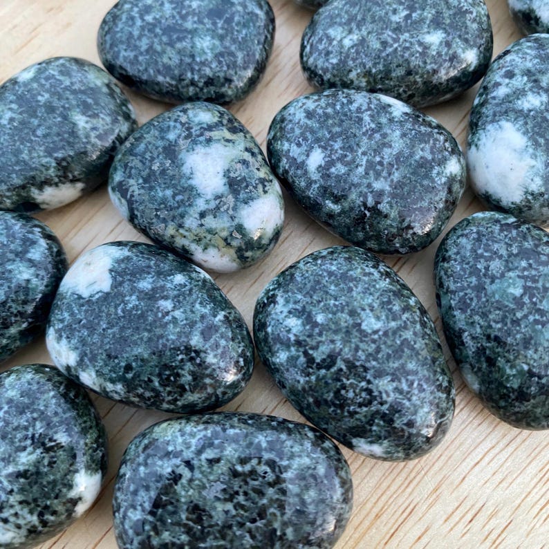 May include: A collection of smooth, oval-shaped stones. The stones are primarily dark green with white and light green marbling. The stones are arranged on a light brown wooden surface. The stones vary in size and shape.