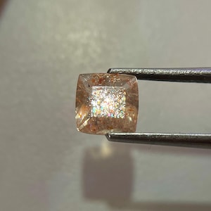 May include: A square-cut sunstone gemstone, held by tweezers. The gem has a peach-colored base with sparkling inclusions that create a shimmering effect. The stone's edges are slightly rounded, and it appears translucent.