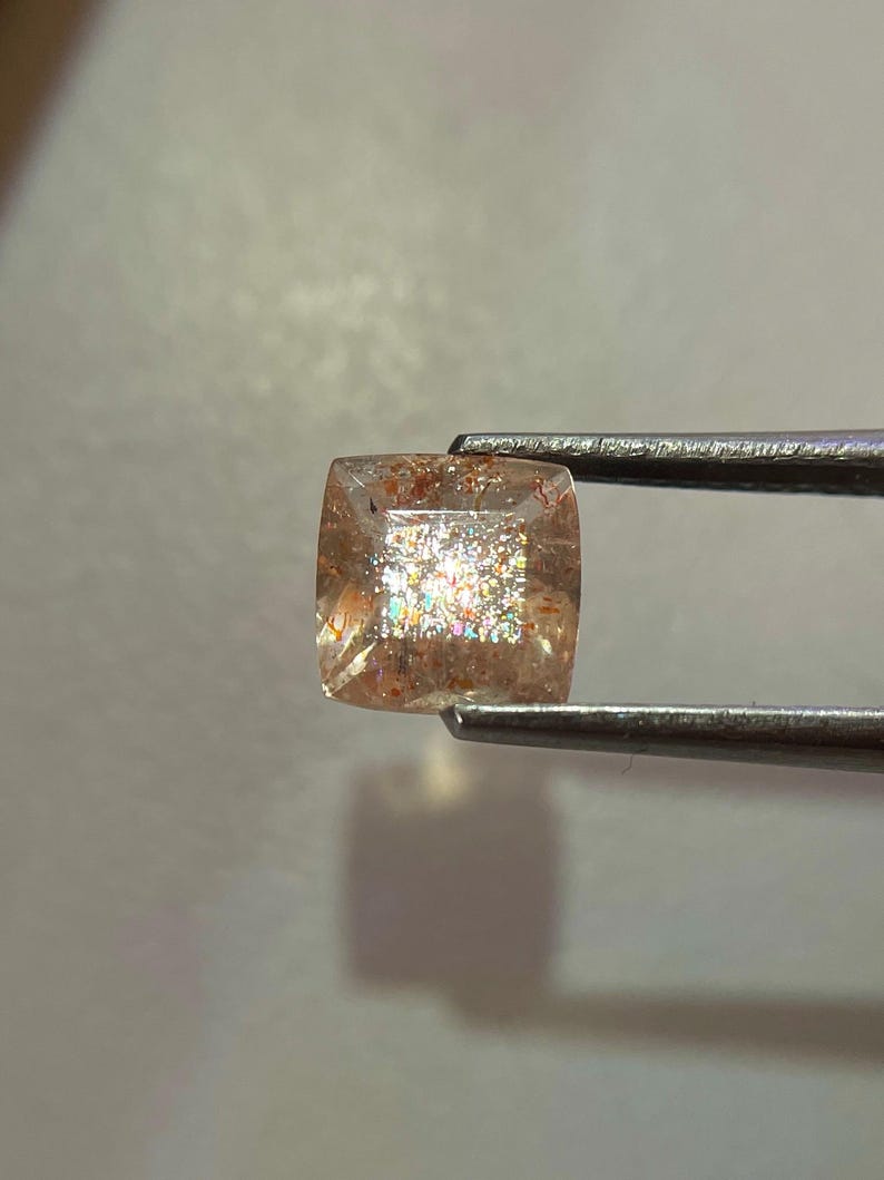 May include: A square-cut sunstone gemstone, held by tweezers. The gem has a peach-colored base with sparkling inclusions that create a shimmering effect. The stone's edges are slightly rounded, and it appears translucent.