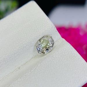 May include: A clear, faceted gemstone with a light green hue, displayed on a white surface.