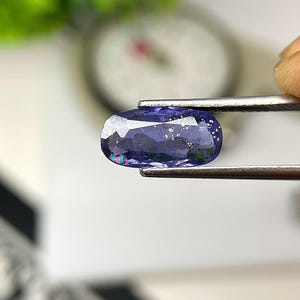 May include: A faceted, oval-shaped, blue-violet gemstone is held by tweezers. The gem has internal inclusions and a subtle play of colours. The background is blurred, with a hint of green and white.