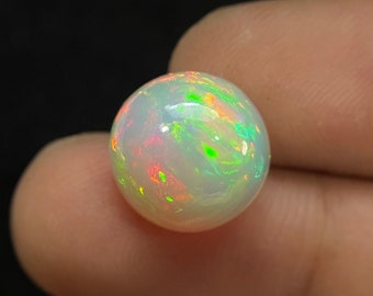 Puede incluir: Una piedra preciosa de ópalo redonda y blanca con destellos iridiscentes verdes y rojos.