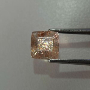 May include: A square-cut gemstone held by tweezers. The gem has a translucent, peach-colored base with internal inclusions and a shimmering, iridescent effect. The gem is likely a sunstone, a type of feldspar.