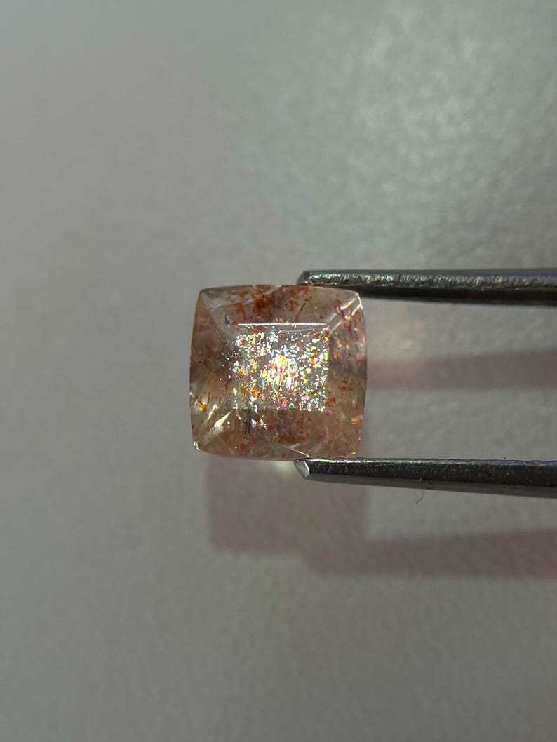 May include: A square-cut gemstone held by tweezers. The gem has a translucent, peach-colored base with internal inclusions and a shimmering, iridescent effect. The gem is likely a sunstone, a type of feldspar.