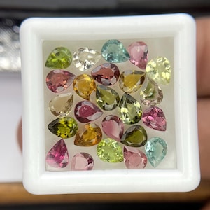 May include: A collection of pear-shaped gemstones in a white square container. The gems display a variety of colors, including pink, green, yellow, and clear. The gemstones are arranged closely together, filling the container.