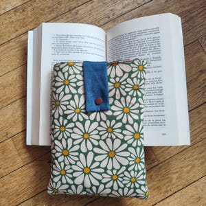 Peut inclure: Une housse de livre rectangulaire en tissu avec un motif répétitif de marguerites blanches à cœur jaune sur fond vert foncé. La housse possède une patte de fermeture en tissu bleu style jean, fixée par un bouton-pression circulaire rouge foncé.