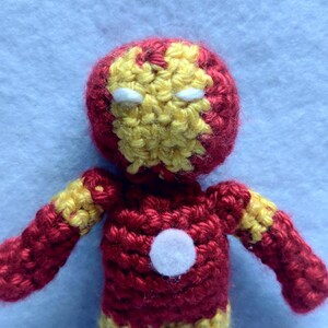 May include: A crocheted plush toy of Iron Man, a superhero from Marvel Comics. The toy is red and yellow with a white face mask and a white circle on the chest.