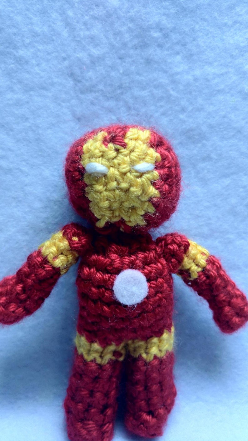 May include: A crocheted plush toy of Iron Man, a superhero from Marvel Comics. The toy is red and yellow with a white face mask and a white circle on the chest.