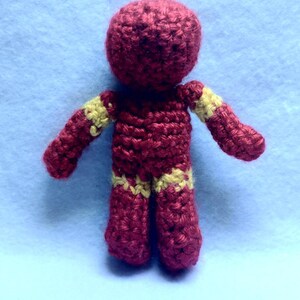 May include: A crocheted red and yellow superhero figurine. The figurine is standing with its arms outstretched.