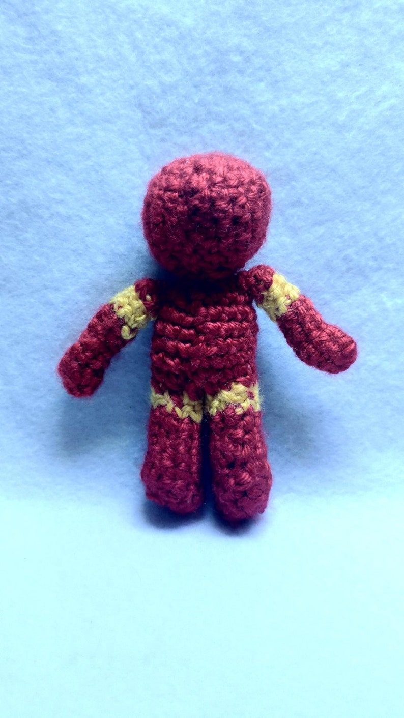 May include: A crocheted red and yellow superhero figurine. The figurine is standing with its arms outstretched.