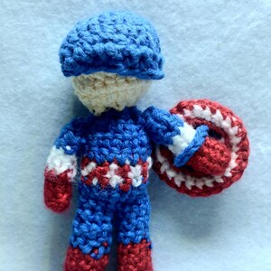 May include: A crocheted toy figure of Captain America, wearing a blue hat, blue and red suit, and holding a red, white, and blue shield.
