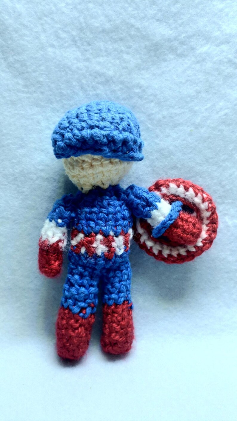 May include: A crocheted toy figure of Captain America, wearing a blue hat, blue and red suit, and holding a red, white, and blue shield.