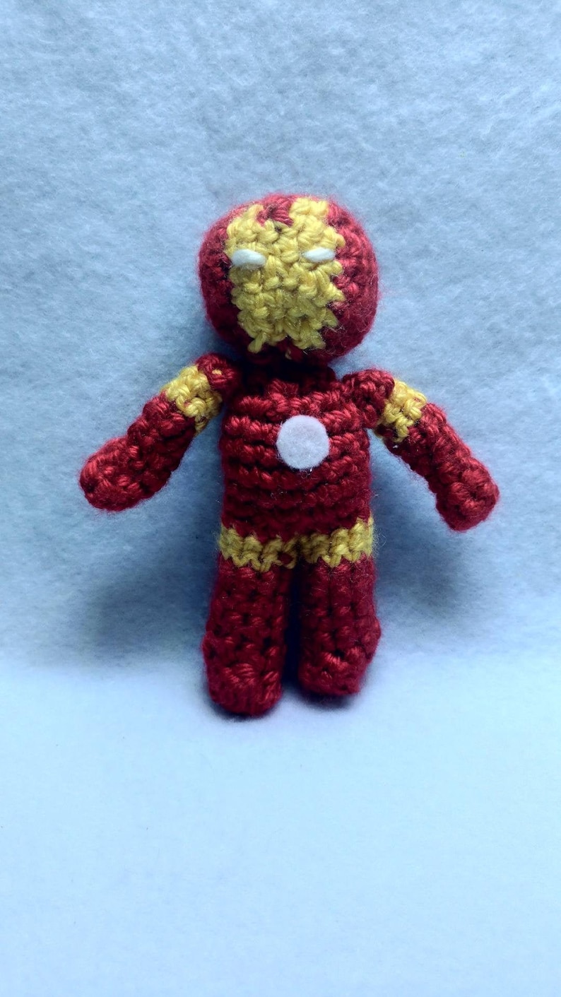 May include: A crocheted toy figure of Iron Man, a superhero from Marvel Comics. The figure is red and yellow with a white circle on the chest. The figure has a crocheted face with yellow eyes and a red mouth.