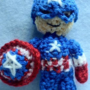 May include: A crocheted amigurumi toy of Captain America. The toy is wearing a blue and white crocheted costume with a red star on the chest. The toy is holding a red, white, and blue crocheted shield.