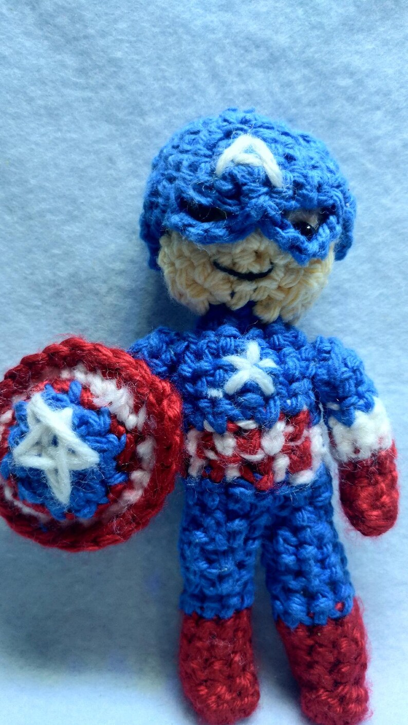 May include: A crocheted amigurumi toy of Captain America. The toy is wearing a blue and white crocheted costume with a red star on the chest. The toy is holding a red, white, and blue crocheted shield.