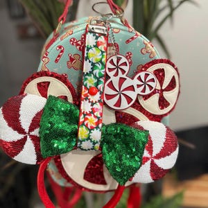 May include: A mint green handbag featuring a candy cane and gingerbread man design. The bag is adorned with a candy-themed keyring and a red and white peppermint-themed headband with green sequin bows and peppermint ear accents.
