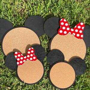 May include: Two corkboard mouse head shapes with black ears and red bows with white polka dots. The corkboard is brown and the ears are black.