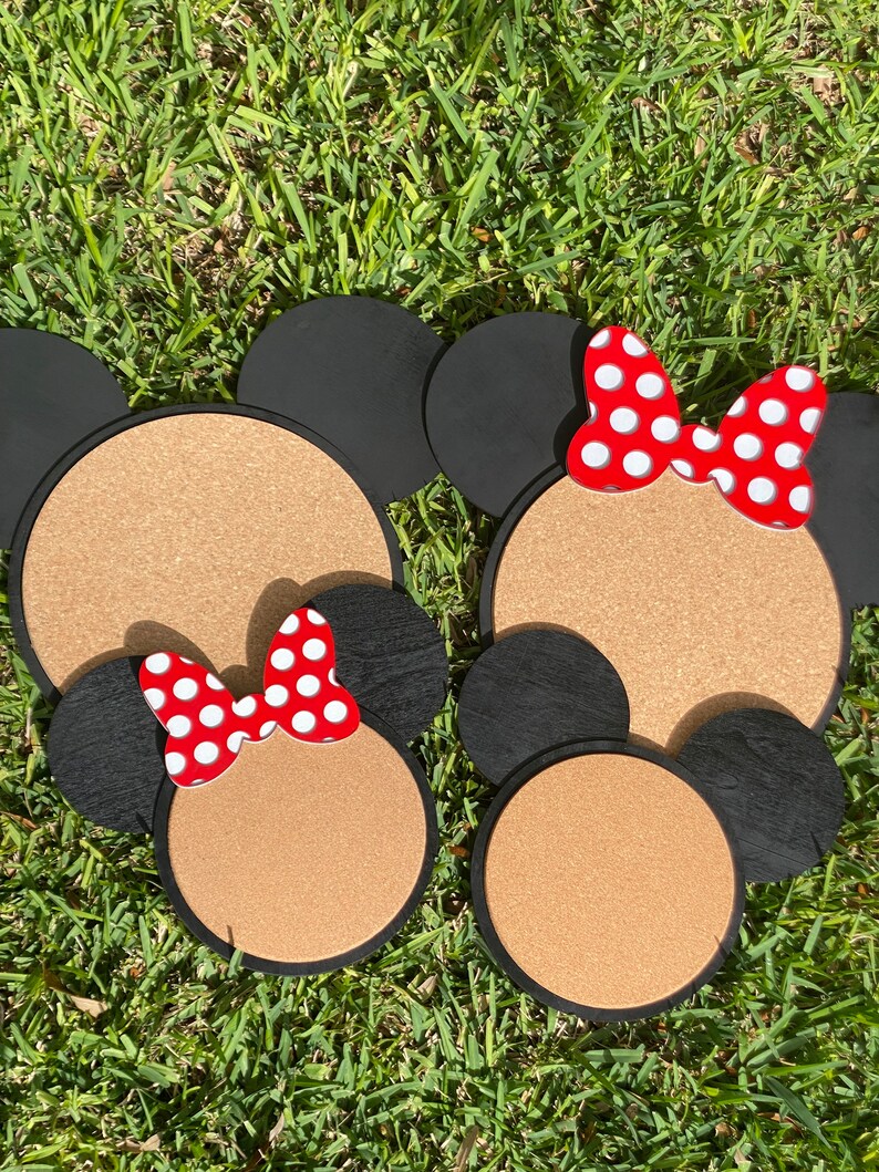May include: Two corkboard mouse head shapes with black ears and red bows with white polka dots. The corkboard is brown and the ears are black.
