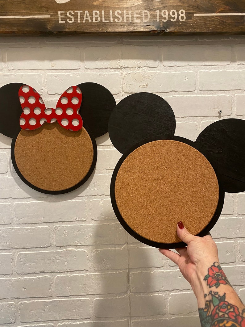 May include: Two corkboard Mickey Mouse head silhouettes, one with a red and white polka dot bow. The corkboards are black with a brown cork centre.