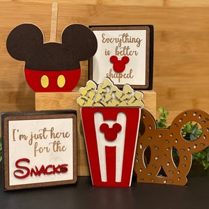 May include: Four wooden signs with a Mickey Mouse theme. The signs feature the text "I'm just here for the snacks", "Everything is better shaped", a Mickey Mouse head, and a popcorn bucket with Mickey Mouse ears. The signs are decorated with red, white, and brown paint.