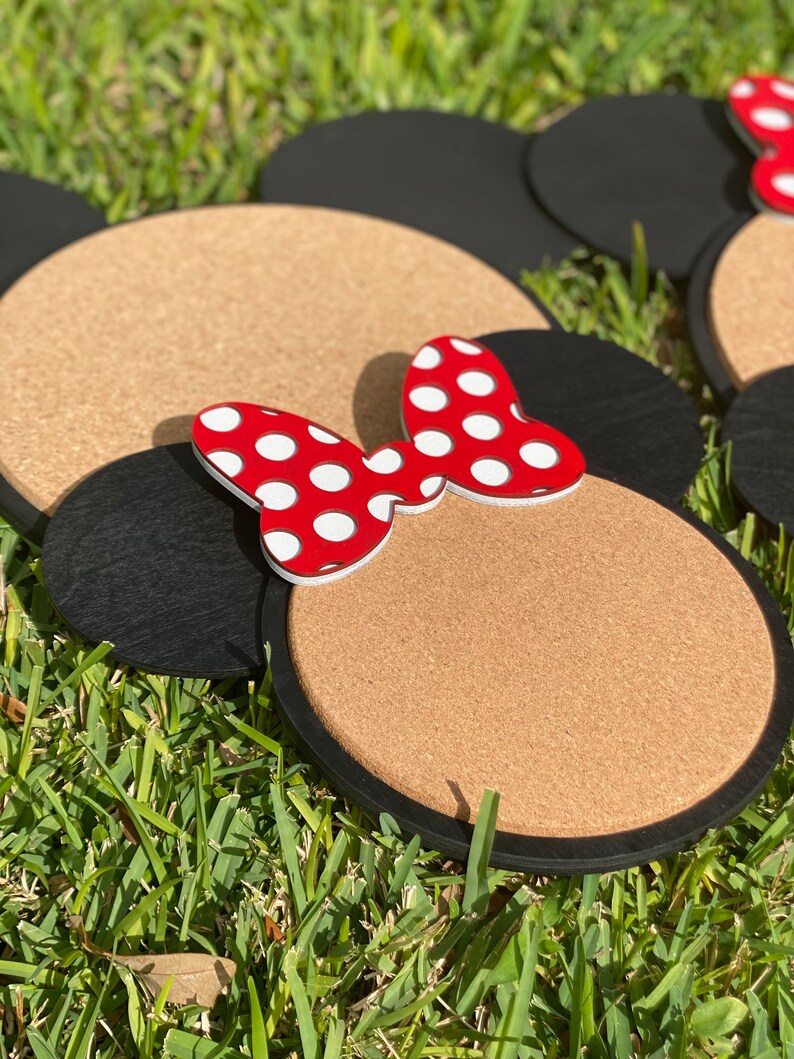 May include: Two corkboard mouse ears with black felt trim and a red bow with white polka dots. The ears are on a green grassy surface.