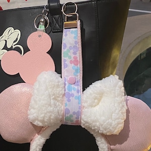 May include: A black tote bag featuring a Minnie Mouse design, a pink Mickey Mouse keychain, and a pastel-coloured wristlet with a button closure. The bag has a red and white patterned interior, and pink mouse ears with a white fluffy bow.