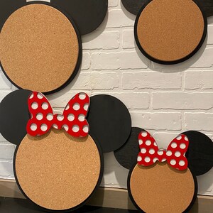 May include: Three corkboard notice boards shaped like Mickey Mouse heads. The largest board has black ears and a brown corkboard face. The middle board has black ears and a brown corkboard face with a red bow with white polka dots. The smallest board has black ears and a brown corkboard face with a red bow with white polka dots.