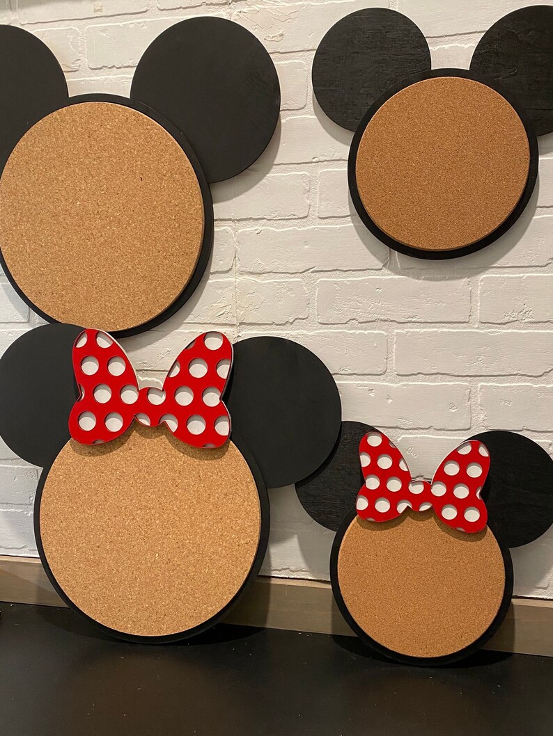 May include: Three corkboard notice boards shaped like Mickey Mouse heads. The largest board has black ears and a brown corkboard face. The middle board has black ears and a brown corkboard face with a red bow with white polka dots. The smallest board has black ears and a brown corkboard face with a red bow with white polka dots.