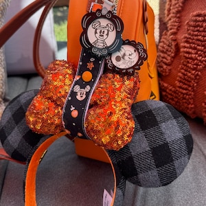May include: Black and grey plaid Minnie Mouse ears headband with an orange bow decorated with orange and gold sequins. The headband is attached to a brown leather bag with a Mickey Mouse charm.