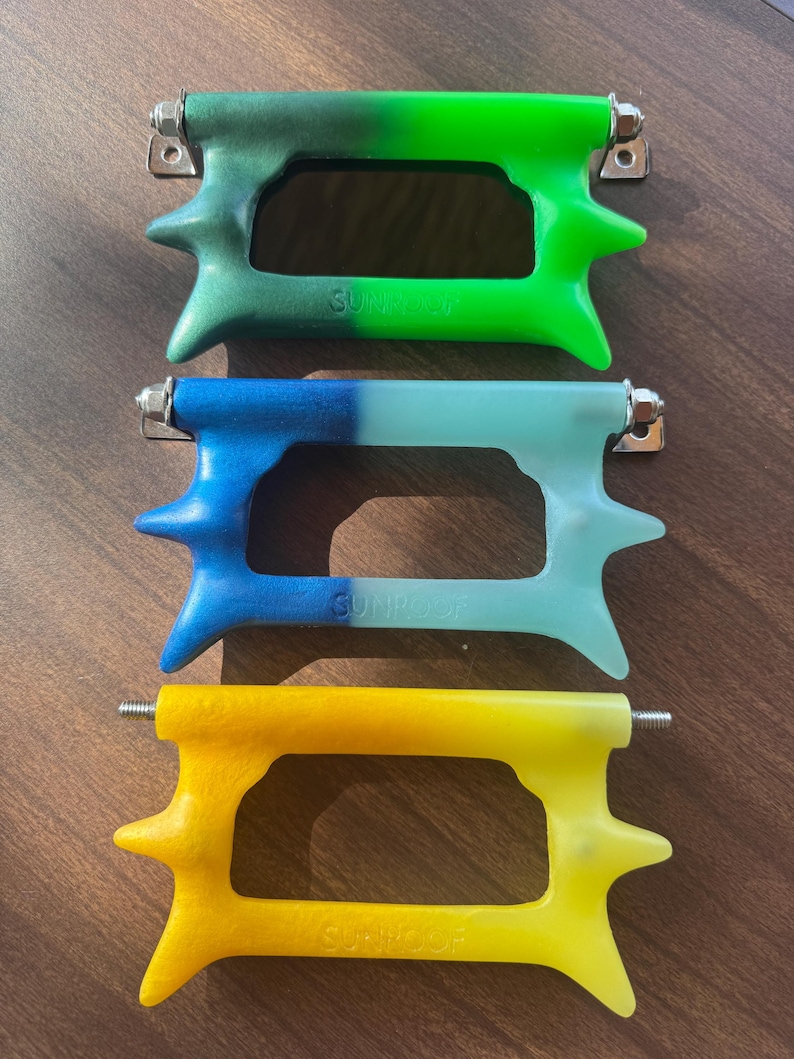 May include: Three colourful, stylised handles with the word "SUNROOF" embossed on each. The handles are green, blue, and yellow, each with a unique gradient effect. They have a distinctive shape with pointed protrusions and metal hardware.