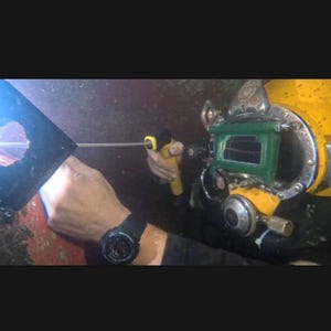 K&ouml;nnte beinhalten: Ein Taucher mit einem gelb-gr&uuml;nen Taucherhelm, der ein gelbes Werkzeug h&auml;lt. Der Taucher tr&auml;gt eine schwarze Uhr. Der Hintergrund ist dunkel und verschwommen, was auf eine Unterwasserumgebung hindeutet.