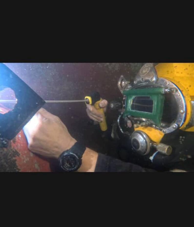 K&ouml;nnte beinhalten: Ein Taucher mit einem gelb-gr&uuml;nen Taucherhelm, der ein gelbes Werkzeug h&auml;lt. Der Taucher tr&auml;gt eine schwarze Uhr. Der Hintergrund ist dunkel und verschwommen, was auf eine Unterwasserumgebung hindeutet.