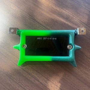 May include: A green and black rectangular device with the text "ANSI Z87-1-K-9-H" printed on the front. The device has metal brackets on each end and is secured with two screws. The device is on a wooden surface.