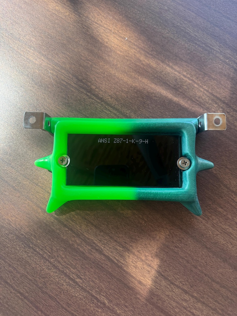 May include: A green and black rectangular device with the text "ANSI Z87-1-K-9-H" printed on the front. The device has metal brackets on each end and is secured with two screws. The device is on a wooden surface.